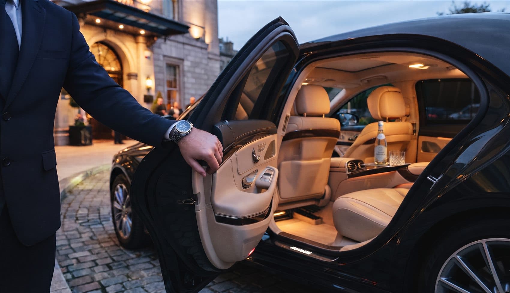 Chauffeur holding the rear door open outside a hotel at dusk